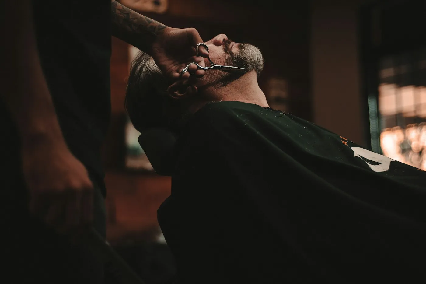 A barber shaping a reclining client’s beard with a straight razor in a dimly lit shop.
