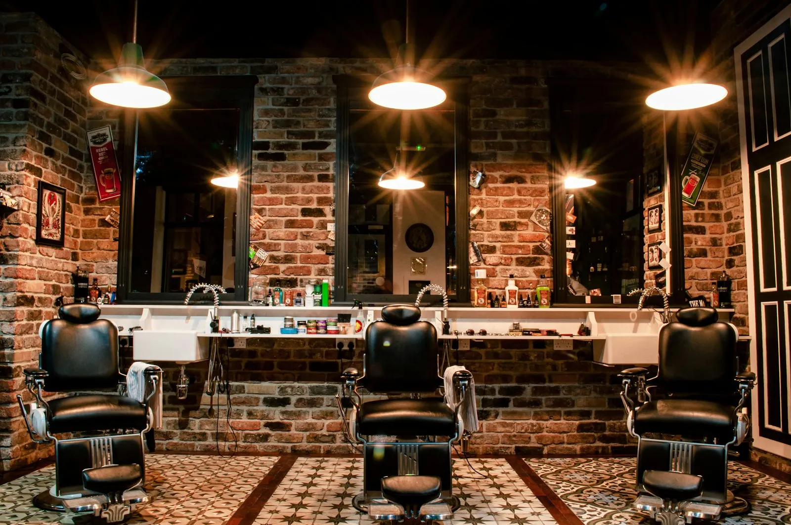 The interior of a modern barbershop with three empty barber chairs lit by warm pendant lights against an exposed brick wall.